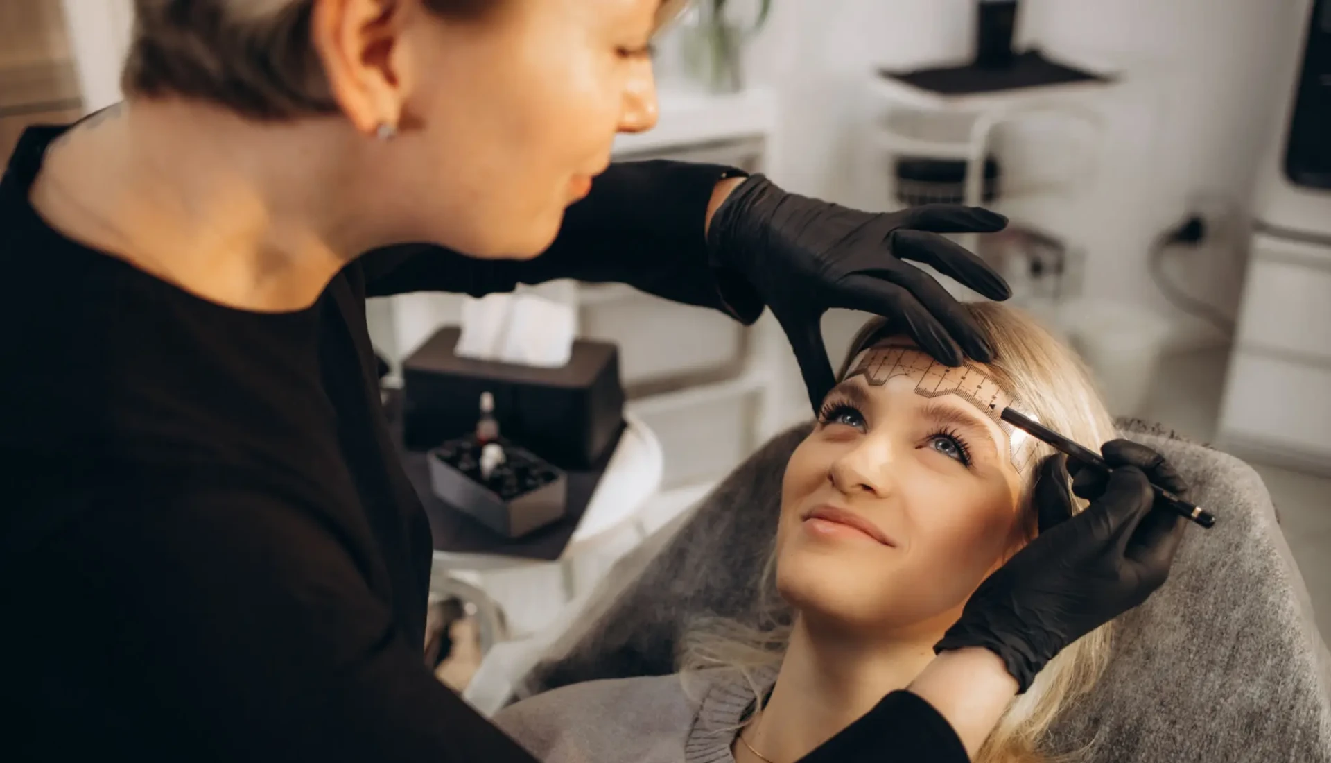 A woman receiving eyebrow shaping treatment from a beautician.
