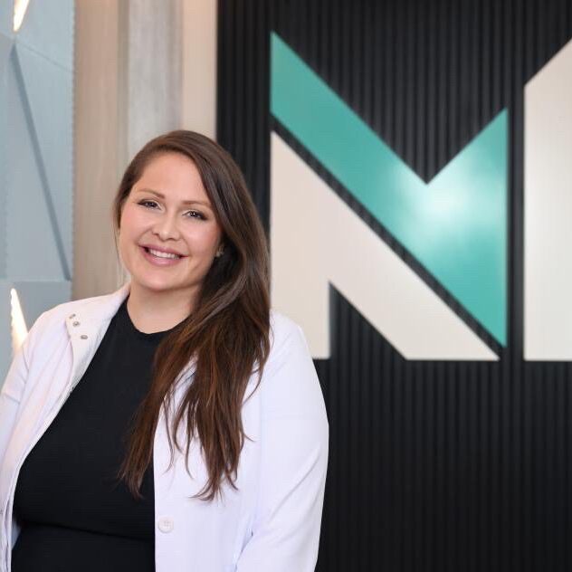 Smiling woman in a white coat standing indoors near a modern logo wall.