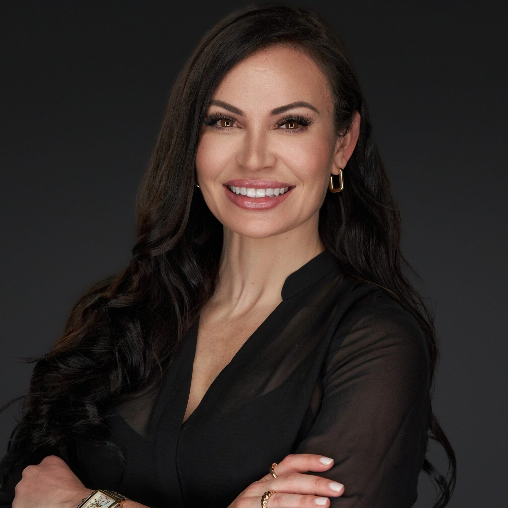 Confident woman with long dark hair in a black blouse, smiling warmly against a dark background.