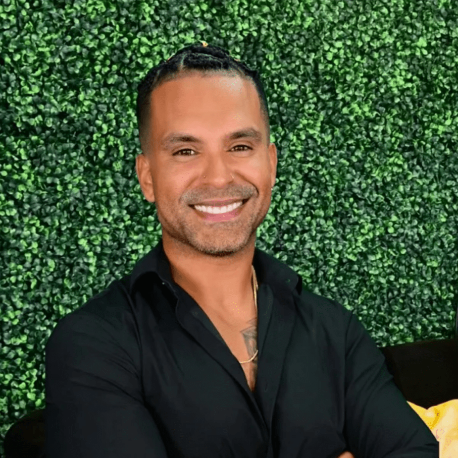 Smiling man in black shirt stands confidently against green foliage.